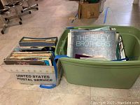 Two boxes filled with vinyl record albums, one green plastic bin and one USPS cardboard box, showing various album covers.
