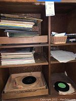 Shelf with stacks of vinyl records and some loose records in paper sleeves, along with a few cassette tapes on a shelf and in a basket.