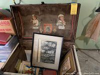 Open antique wooden trunk showing framed black and white and colored artwork inside along with books and papers.