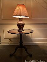 Round dark wood table with scalloped edges and carved details, lamp on top lit with pleated shade