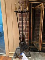 Full view of the black metal fireplace tool stand holding broom, shovel, tongs, and poker with brass handles, positioned in front of a brick fireplace.