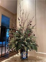 Full view of the tall floral arrangement showing mixed greenery, flowers, twigs, and tall branches in metal container against wall and banister.