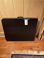 Black folding table folded and standing upright showing the carrying handle and smooth black surface.