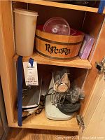 Cabinet shelf showing Sunbeam mixer, Nutrichef food saver, popcorn bucket, and lilac napkins