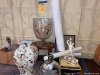 Wide shot showing large etched glass bowl on small wooden stool, floral vase, mercury glass vase, rolled posters, small sculpture on books, and a box with miscellaneous items.