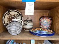 Five assorted ceramic home decor items arranged on wooden shelf: two plates, one pot with lid, one pitcher, one vase.