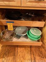 Open kitchen drawer showing cast iron pan, black frying pans, blue and clear rectangular Pyrex dishes, and green Tupperware containers with lids