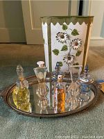 Photo showing all perfume bottles arranged on the oval mirrored tray with the floral metal wastebasket in the background on the floor.