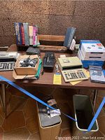 Wide angle photo showing the lot content on table including calculators, electronic organizer, packages of printer paper, reference books on stand, lock box, and other items.