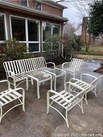 White metal outdoor patio furniture with one couch, four chairs, and two black metal glass-top tables set on a concrete patio in front of a house.