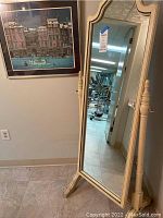 Full length standing painted floor mirror with decorative carved frame and side supports, placed near a framed lithograph painting on the wall.