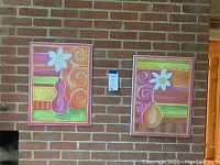 Two colorful abstract floral paintings hung on brick wall, each with white flower and multicolored horizontal striped and swirl background