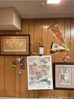 Two signed and framed children themed art prints on wood-paneled wall with vintage travel pennants and assorted sports memorabilia pins and photos below