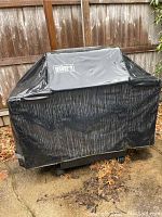Full view of Weber gas grill covered with a black Weber-branded grill cover showing wear and outdoor placement on leaf-strewn concrete patio.