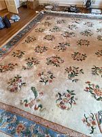 Overview photo showing the full wool rug spread out on wooden floor, beige ground with floral motifs and a blue floral border.
