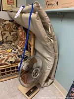 Photo of folded beige wrestling mat leaning against furniture with a blue strap securing it, and a vintage oscillating fan with metal blades on the floor in front.