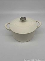 Front angle photo of ivory white enameled cast iron dutch oven with lid on showing both side handles and stainless steel lid knob. Small chip visible on handle.