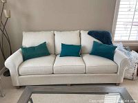 Full frontal view of cream Bassett sofa with three teal accent pillows placed on it. Sofa has rolled arms and three seats with separate cushions.
