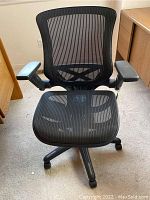 Front view of the black mesh office chair with armrests and caster wheels on a light carpet.