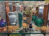 Overview of glassware and decorative beads on top glass shelf including light blue vase, tall clear vase with blue beads, iridescent glass sphere, small artificial plant and gold bead strands.