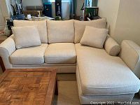 View of beige fabric upholstered convertible couch with chaise section and matching ottoman nearby. Includes three seat cushions, three back cushions, and two extra small cushions.