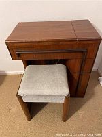 Full view of the wooden sewing machine cabinet with closed top and the cushioned stool in front.