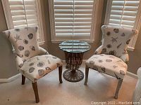 Pair of seashell printed accent chairs with wooden legs and decorative metal loop on back shown near window with blinds. Glass accent table positioned between chairs not included.