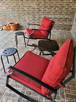 Two rocking chairs with red cushions, black metal frame, a square side table with black glass top, and two small black metal stools with holes in round seats on a brick patio.