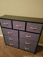 Angled front view of 9 drawer dresser with purple-gray fabric drawers and wooden handles. Black wood frame and top.