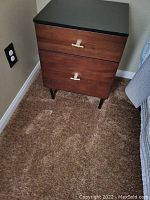 Front view of a two drawer nightstand against a wall with carpet flooring. Shows black top, wood drawers with gold-tone handles, tapered legs.