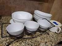 White porcelain bowls, medium size, round shape stacked in front, four long oval white porcelain dishes stacked behind, three small round white bowls displayed on a black metal holder, and a white cup with a blue lid beside them on granite counter.