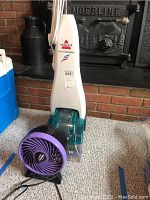 Bissell Quick Steamer 655 and purple fan together on carpet in front of a brick fireplace. Shows front and side of steamer and fan.