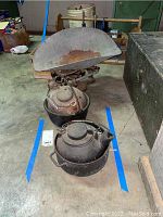 Photo showing two cast iron kettles/pots with coil handles placed inside a larger pot, vintage metal balance scale behind them showing rust and wear.