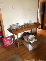 Wooden table with stacked ceramic plates, glassware, and a cardboard box containing holiday plates on floor.