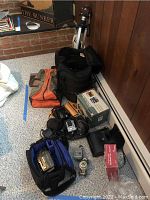 Overview of lot showing the camera tripod, electronic devices in cases and boxes, various bags containing cameras and accessories, and other items arranged on floor.
