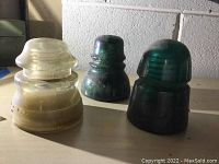 Three glass electrical insulators placed on a surface against a cinder block wall, one clear and two green, showing condition and size differences.