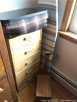 Cardboard storage drawers with five drawers and cream finish, topped with an ironing board with dark plaid fabric cover, with wooden stool in front.
