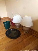 Photo showing two matching table lamps with cream ceramic bases and white shades, a small green trashcan, a small wooden stool, and a round wicker tray with glass center, all placed on hardwood floor against a beige wall.