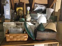 Various lighting items with some storage and decor, including two oil lamps with clear hurricane glass shades, a lamp with shade (green base and cream fabric shade), a plastic lantern, and a wooden box with miniature tools.