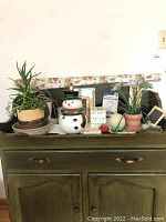 Photo showing potted plants, a ceramic snowman container, several picture frames, and wrapping paper on top of a green wooden cabinet.