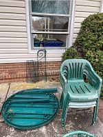 Stacked green plastic chairs with curved armrests, a round disassembled table with visible metal frame from the underside, and a green metal trellis leaning against a wall outdoors on a concrete surface.
