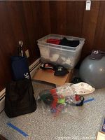 Overview photo showing workout mat rolled up, exercise ball, plastic tote with towels, red boxing gloves, cardboard box, and black mesh bag on floor.
