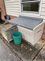 Wide shot showing the large Suncast plastic outdoor storage chest beside the smaller weathered storage box and a green bucket.