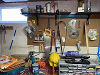 Workbench and pegboard wall stocked with assorted hand tools including wire cutters, hand saws, multiple items hung on pegboard, wooden ruler, yellow hard hat, some shelving storage containers and paints on shelf above.