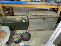 Two wood trunks placed side by side under a workbench cluttered with various tools. Items including two old metal kettles and a rusty metal scoop sit on the floor near the trunks.