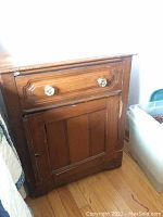 Vintage wooden nightstand with one drawer and one door, showing scratches and damage, including missing hinge on top of door and door edge damage.