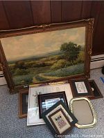 Photo showing a large framed landscape painting with several smaller frames and prints stacked below