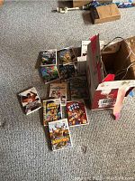 Overview of multiple Wii game cases arranged on carpet near torn cardboard box with cables.