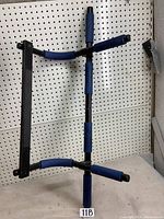 Front view of black metal abdominal workout bar with blue foam grips, positioned against a pegboard wall.