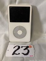 Front view of white Apple iPod Classic showing screen, click wheel, and part of white casing with lot tag number 23